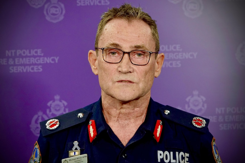 A man in a NT police uniform wearing glasses looks seriously at the camera. The background is purple.