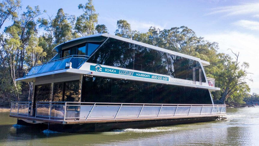 A large double-storey houseboat with Echuca Luxury Houseboats signage sails along a brown river lined with gum trees.