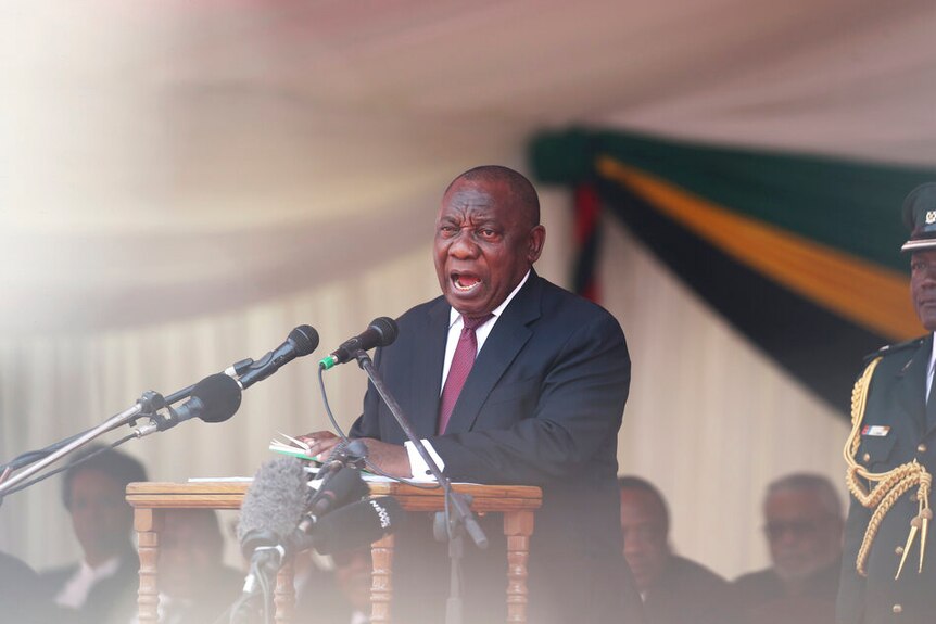 South African President Cyril Ramaphosa stands behind a wooden lectern and two microphones as he makes a speech.