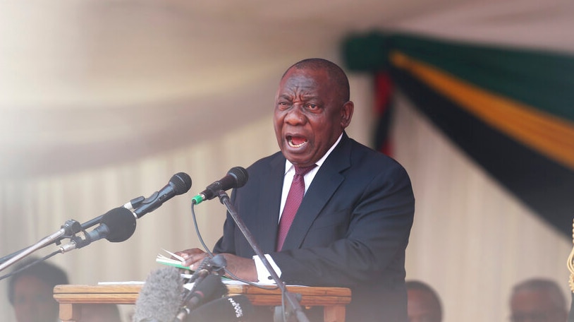 South African President Cyril Ramaphosa stands behind a wooden lectern and two microphones as he makes a speech.
