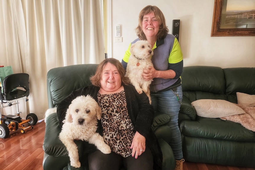 A woman sits on a couch next to a dog, while another woman stands near her holding another dog. Both smile.