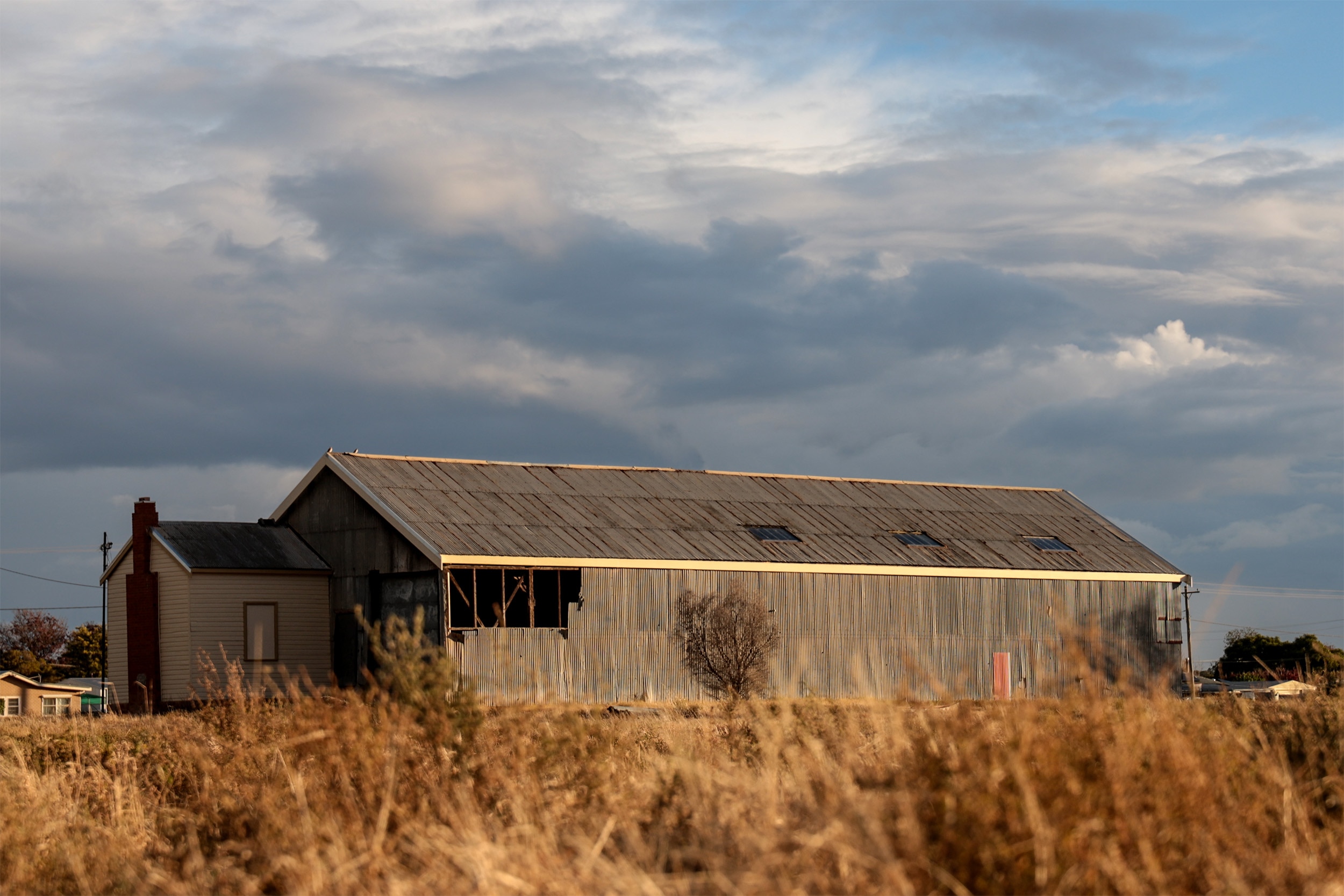 a shed in a state of disrepair, with some tin sheets missing. Yellowed vegetation in front and clouds above.