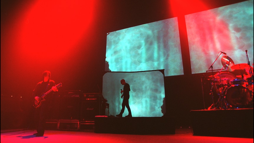 Shadowy figures perform on a stage bathed in red light. Three video screens project a green pattern.
