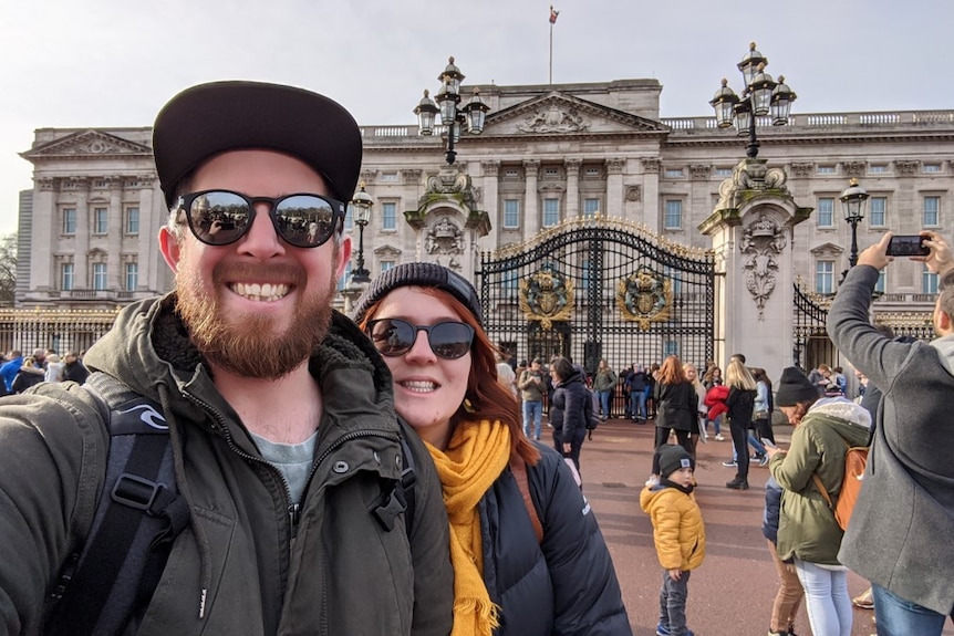 A man with a beard smiles, a woman behind him. They stand outside Buckingham Palace.