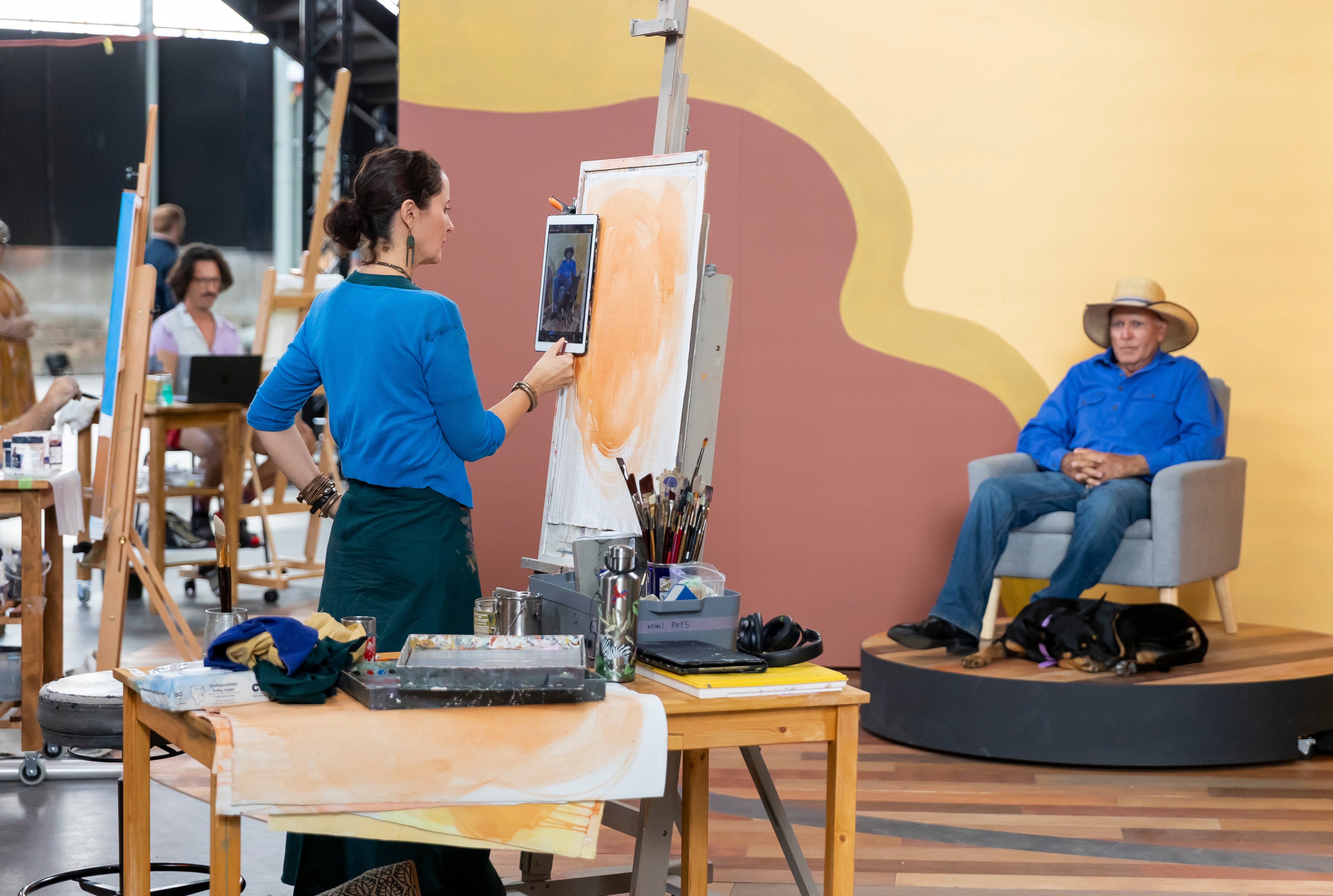 An artist looking at a canvas and her subjects, a man and dog.