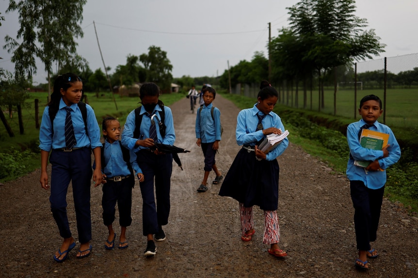 A group of students of different ages all in blue school uniforms walk along a dirt road with yards on either side