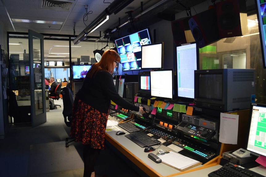 Emma Williams stands in front of a multiple screens in a busy news room.