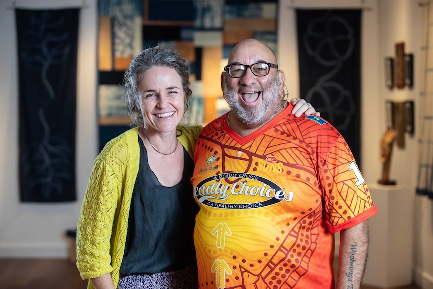 Woman and man posing hugging smiling at camera. Man wearing bright Deadly Choices Gold Coast Suns shirt