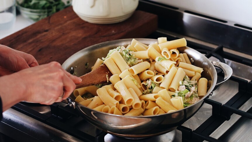 A close-up shows a hand stirring a full pan of tuna pasta with zucchini, basil and rigatoni pasta.