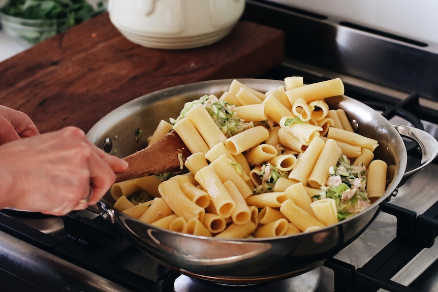 A close-up shows a hand stirring a full pan of tuna pasta with zucchini, basil and rigatoni pasta.