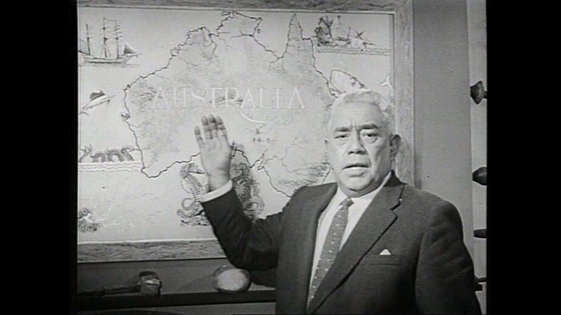 A 1960s black and white photo of an elderly Aboriginal man in a suit, standing in front of a map of Australia