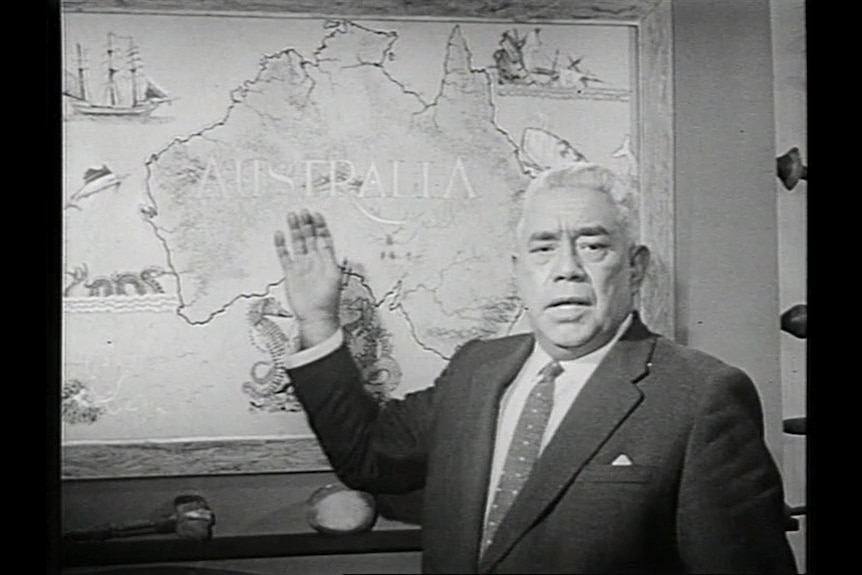 A 1960s black and white photo of an older Aboriginal man in a suit standing in front of a map of Australia