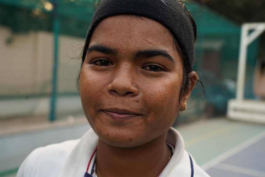 A close up of a woman with brown eyes and wearing a black headband and white polo shirt standing in cricket nets
