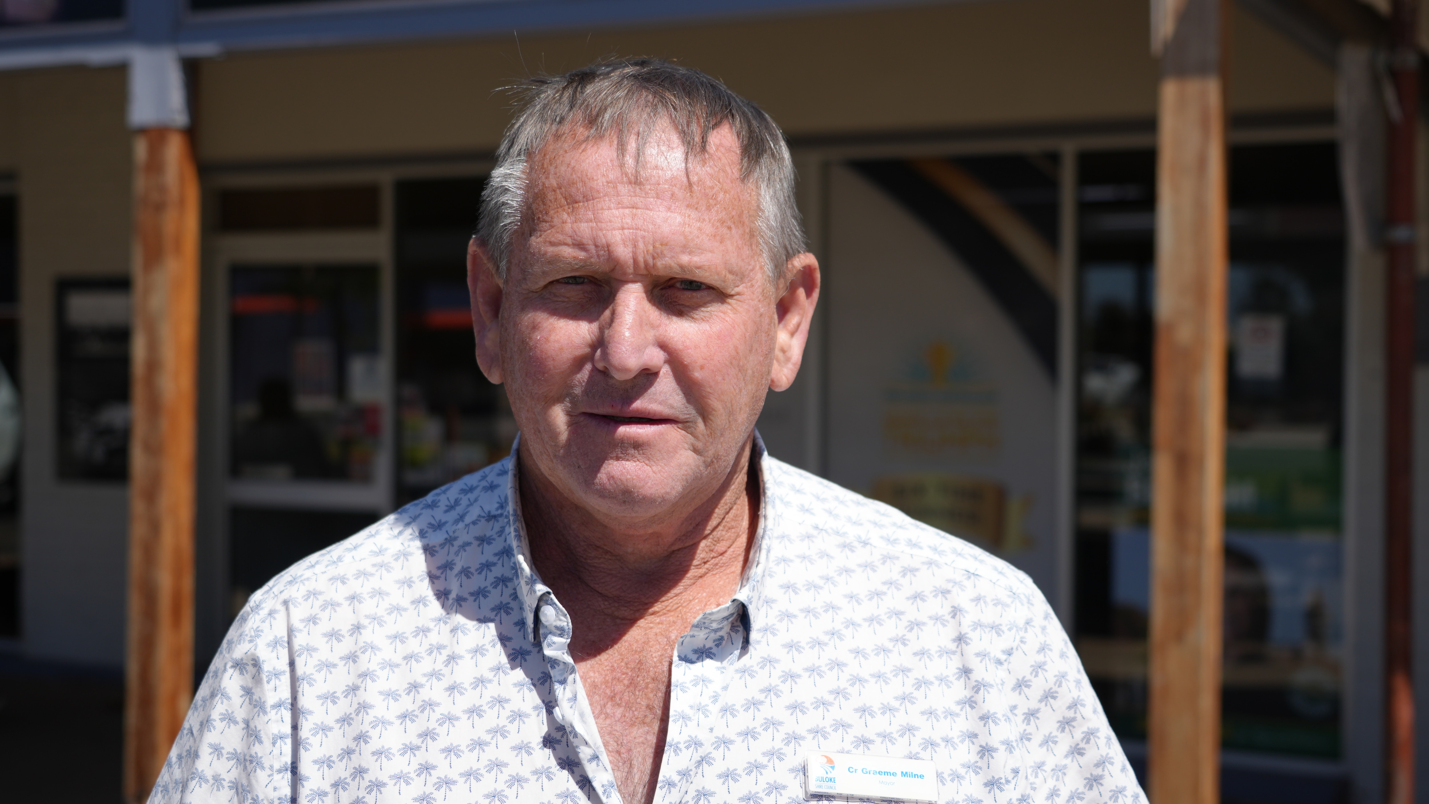 Middle aged man with a white patterened button up shirt stands in sun outside cafe building, serious expression
