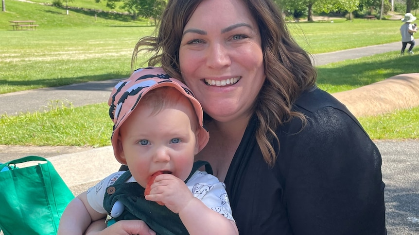 Renee laughs and hugs her baby son Ned while sitting on a picnic blanket outdoors on a sunny day.