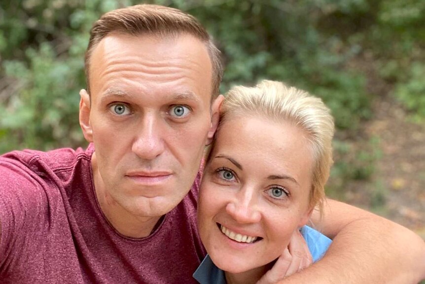 Navalny in a red shirt next to his wife with blonde hair in blue shirt. He has his arm around her with greenery in background.