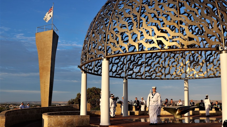 Navy personnel stand to attention under a copper-coloured steel dome atop round poles at the site of the HMAS Sydney II Memorial