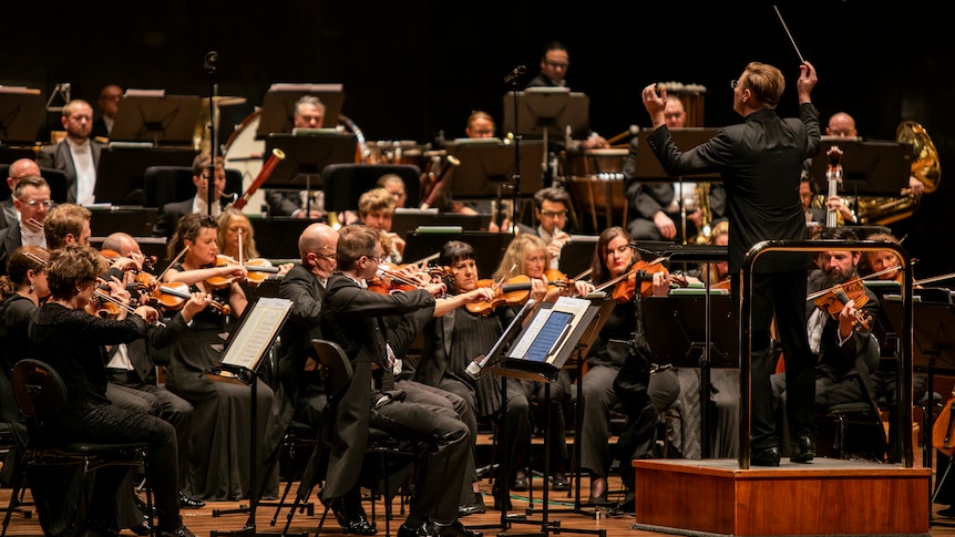 The Melbourne Symphony Orchestra perform on stage. Ben Northey conducts up the front with his baton raised.