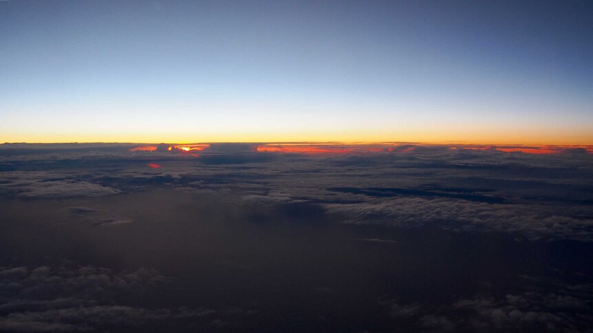 Over patchy, fluffy clouds, a sunrise can be seen on the horizon with deep blue sky above.
