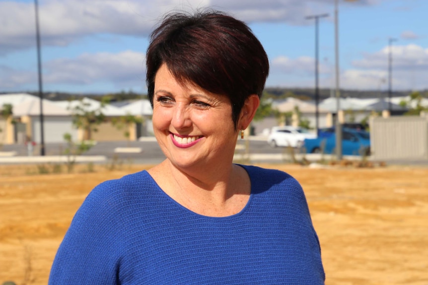 A smiling Alyssa Hayden stands outdoors in front of a housing estate wearing a blue top.