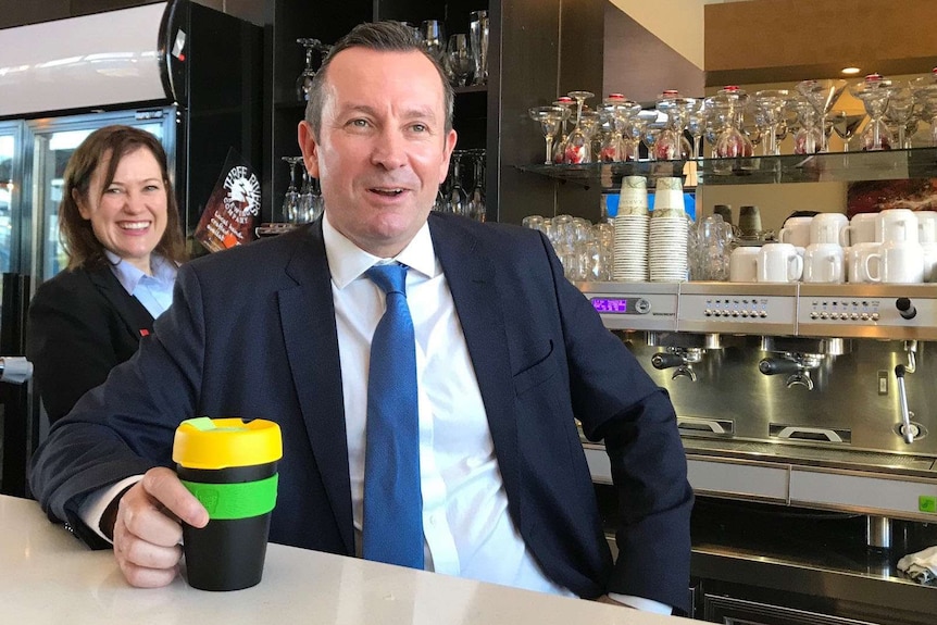 Premier Mark McGowan in a cafe holding a reusable coffee cup, with Tania Lawrence behind him.