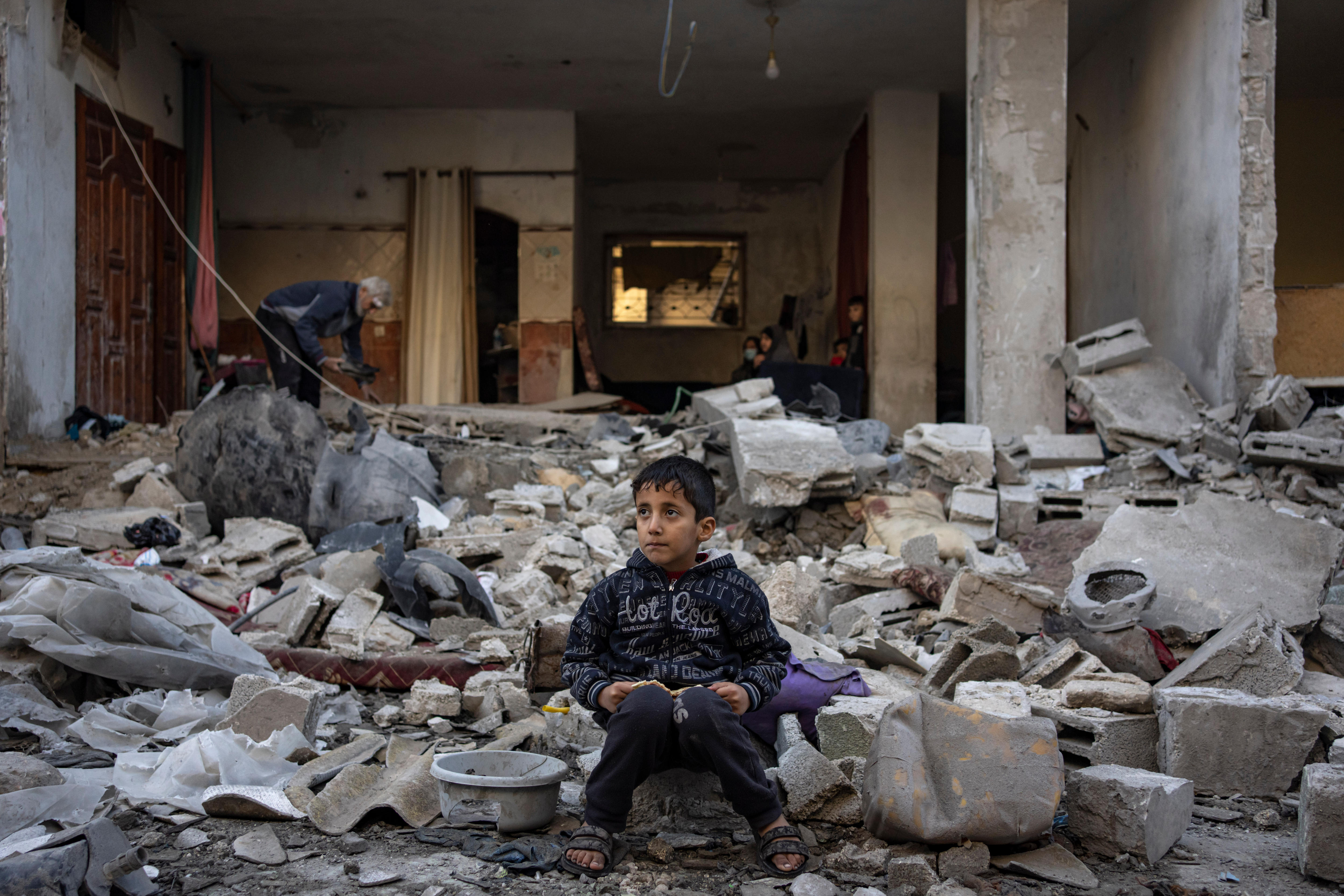 A wide shot of a boy sitting on rubble outside a destroyed building.