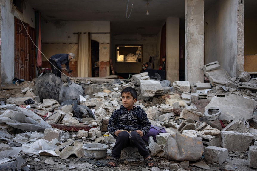 A wide shot of a boy sitting on rubble outside a destroyed building.