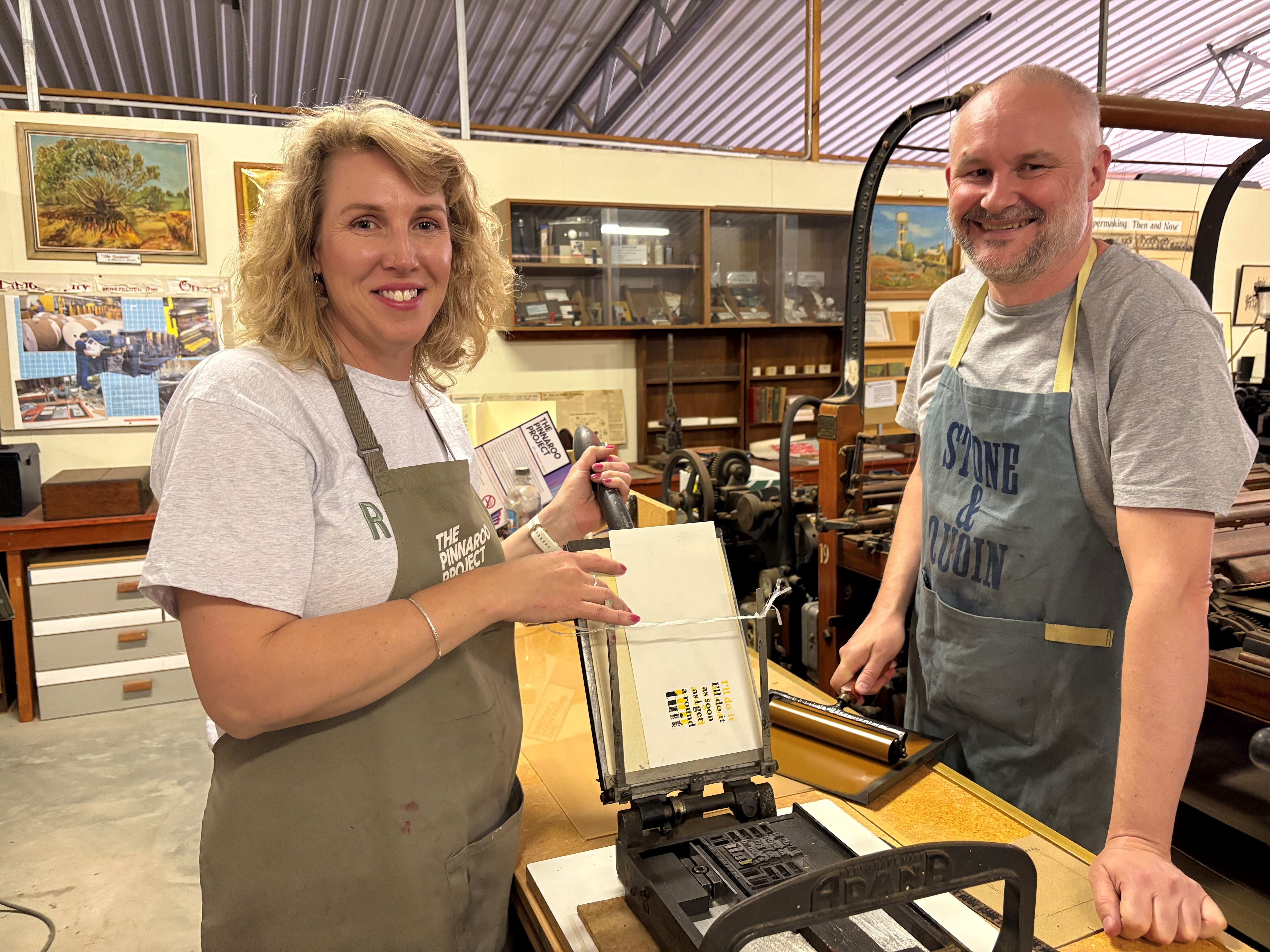 A woman with curly blonde hair and a balding man standing either side of an old, manual printing machine