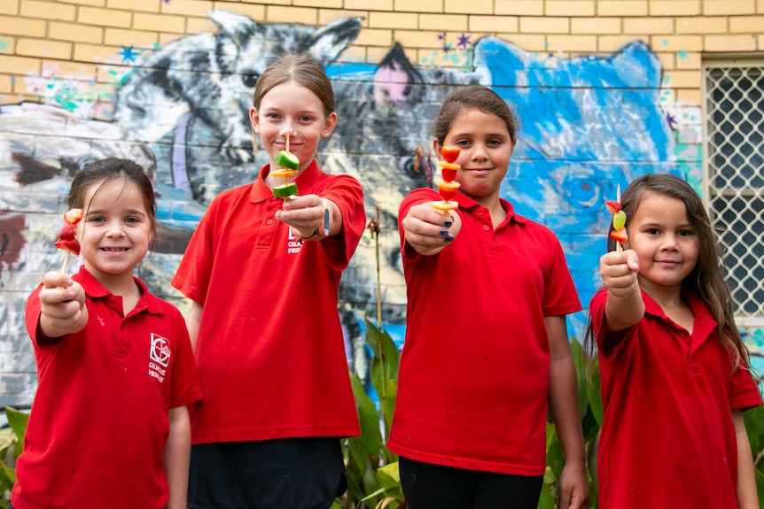 Four young girls in red school shirts hold out fruit skewers.