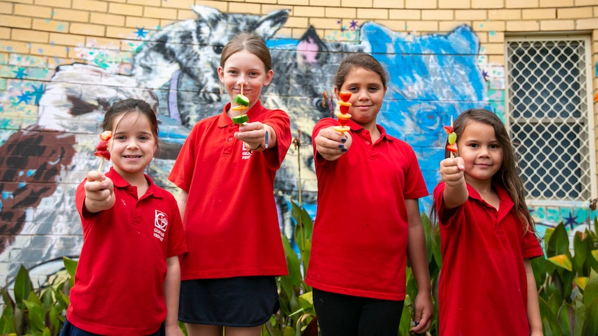 Four young girls in red school shirts hold out fruit skewers.