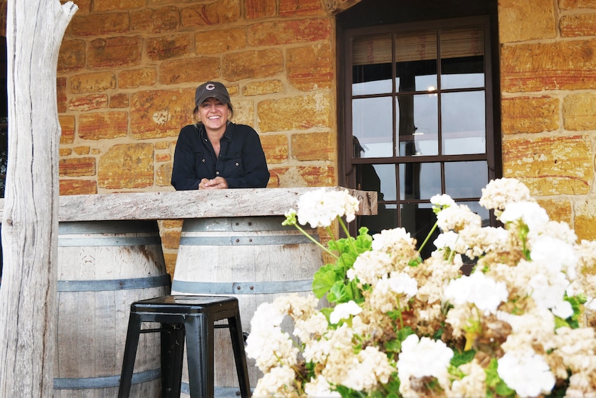 Woman leaning on wooden table outside a stone hut smiling.