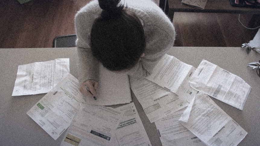 Woman sits with her head in her hands at a table surrounded by bills