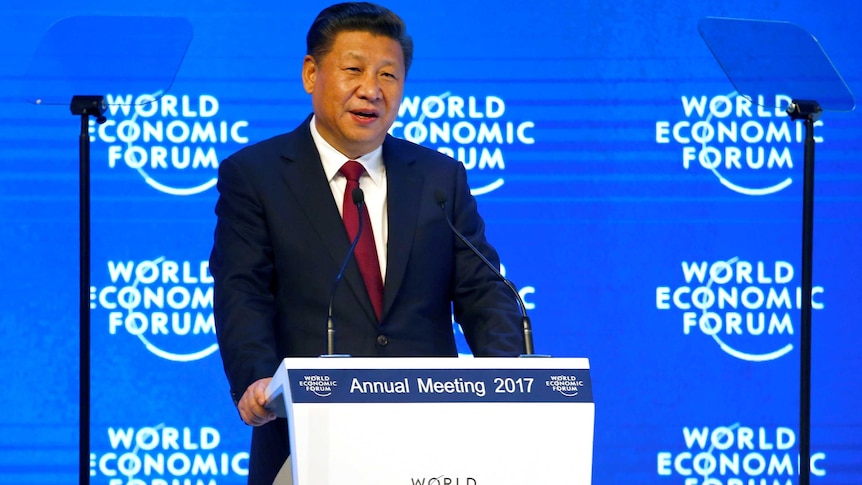 A middle-aged Chinese man in a dark suit stands at podium in front of blue and white screen.