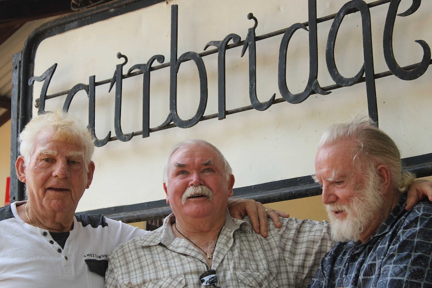 Three men with grey hair stand in front of a sign that says "Fairbridge". They have their arms around each others shoulders.
