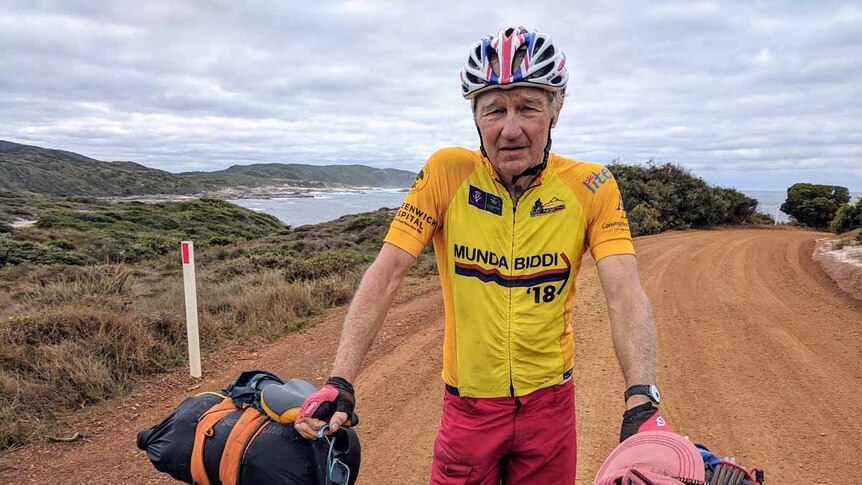 An older man standing on a dirt road with a bicycle, with the ocean in the background.