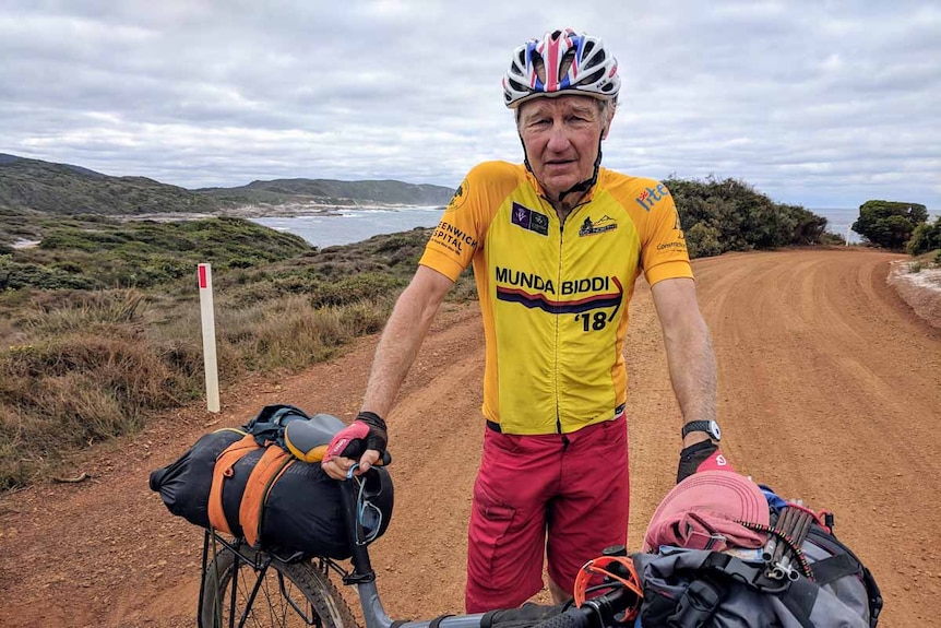 An older man standing on a dirt road with a bicycle, with the ocean in the background.