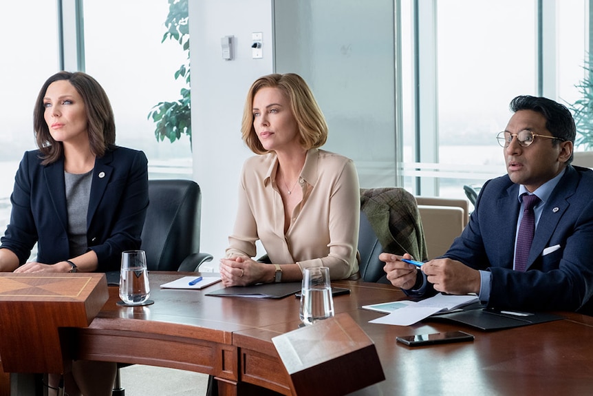 Colour still of June Diane Raphael, Charlize Theron and Ravi Patel seated at wooden table in an office in 2019 film Long Shot.