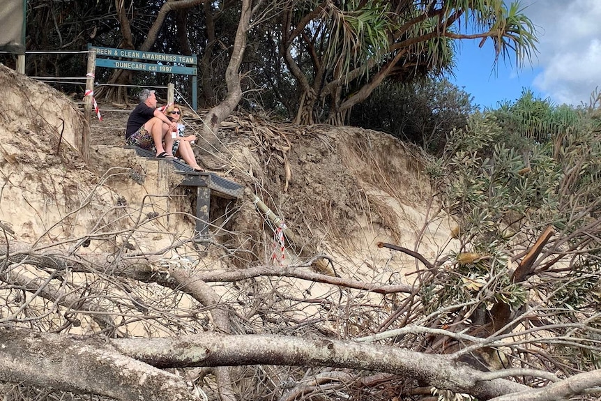 A man and woman sit on a walkway above a badly eroded beach at Byron Bay, and below a Dunecare sign.
