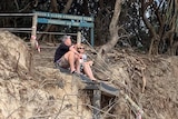 A man and woman sit on a walkway above a badly eroded beach at Byron Bay, and below a Dunecare sign.