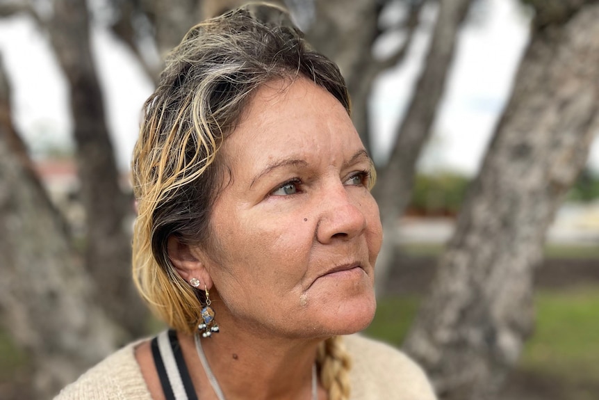 Close up of a woman with blonde pony tailed hair and necklaces sitting on a park bench