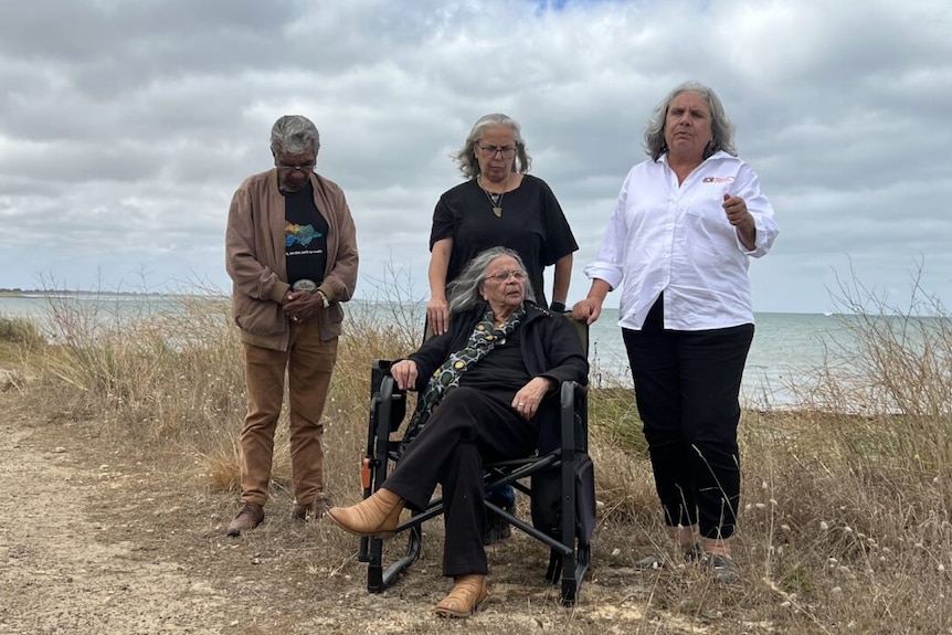 Four older Indigenous people looking solemn as they stand near a body of water on an overcast day.