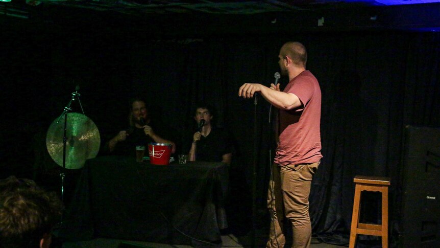 A white man in a maroon shirt performs stand-up, behind him are two white men at a table with a gong speaking during his set.