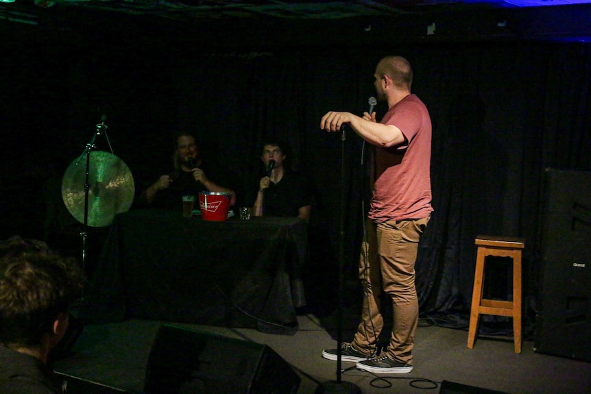 A white man in a maroon shirt performs stand-up, behind him are two white men at a table with a gong speaking during his set.