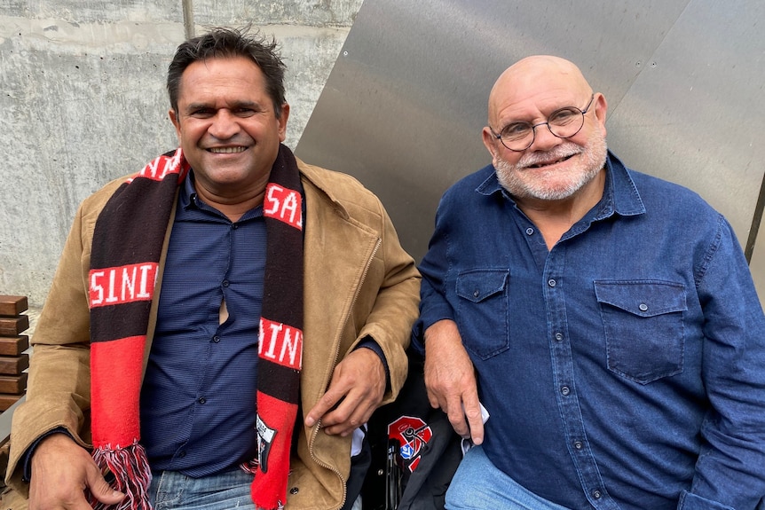 Nicky Winmar and Robert Muir sit and watch an AFL game together.