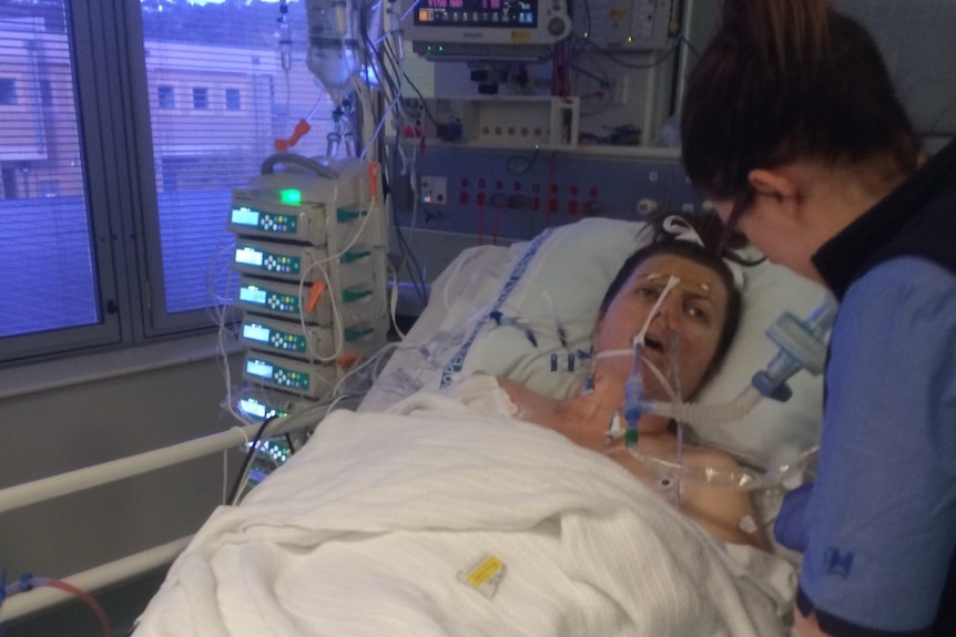 A woman on a ventilator in a hospital bed looks at a female health care worker. 