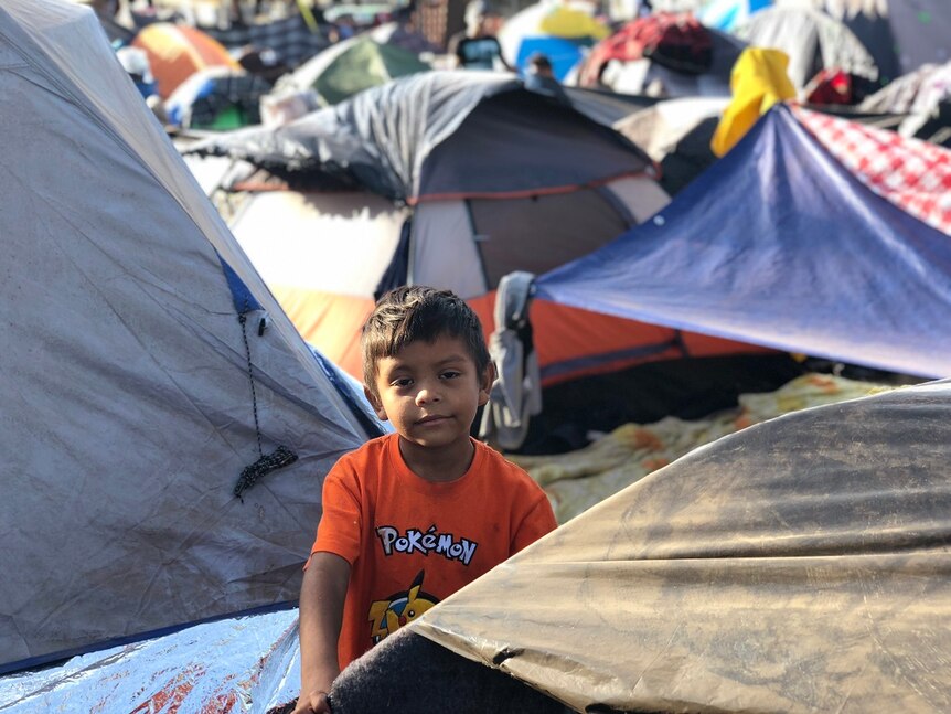 A young boy grips the frame of a tent and looks at the camera while surrounding by densely-packed makeshift accommodation.