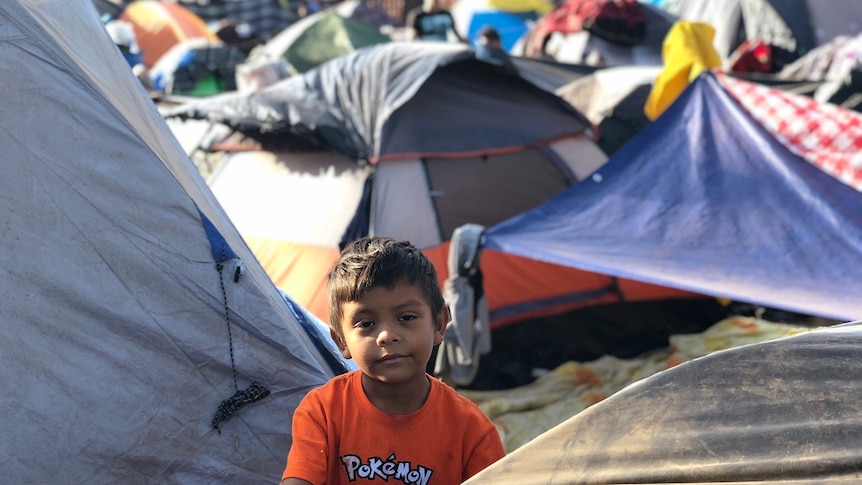A young boy grips the frame of a tent and looks at the camera while surrounding by densely-packed makeshift accommodation.