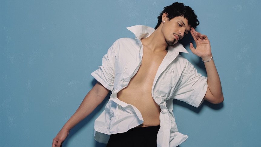 Omar Apollo lays on a blue background with one hand to his head and his white shirt unbuttoned