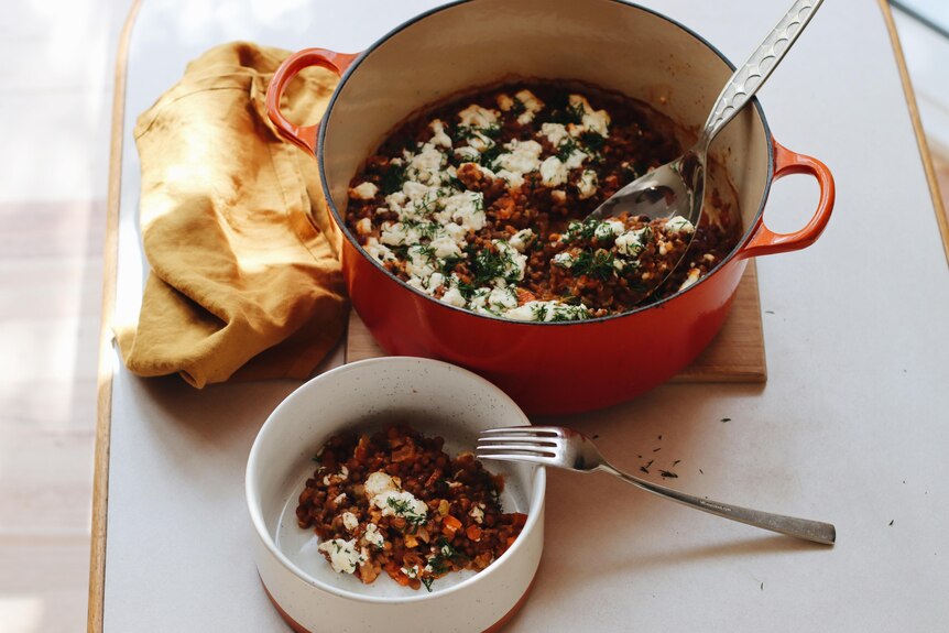 A ladel rests in a Dutch-oven pot filled with lentils and topped with feta cheese and herbs, next to a serving in a bowl.