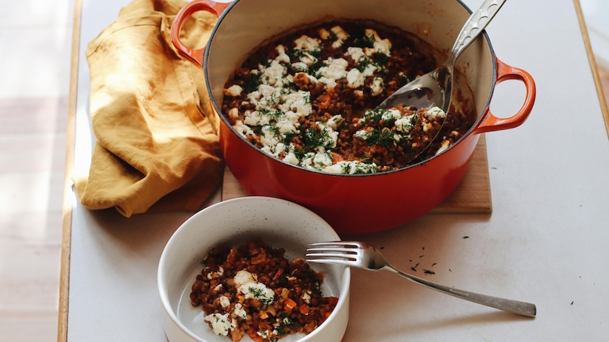 A ladel rests in a Dutch-oven pot filled with lentils and topped with feta cheese and herbs, next to a serving in a bowl.